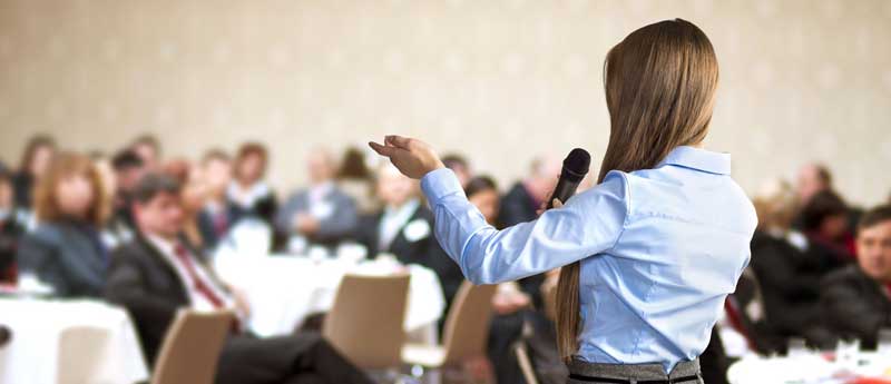 a woman wearing light blue long sleeve speaks in front of the people