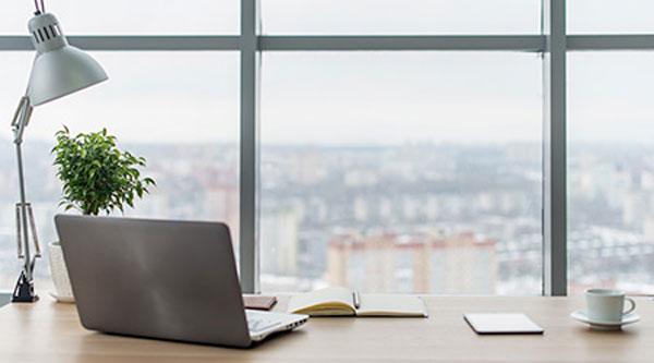 a laptop and papers on a desk with the background of a city through the window.